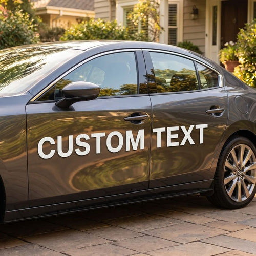 A dark gray sedan parked outdoors on a stone driveway in front of a house. Large white text reading 'CUSTOM TEXT' is printed across the side of the car door.